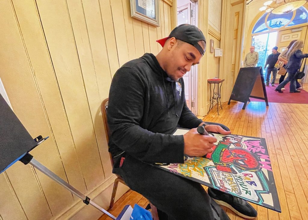 photo of a man seated in a well-lit entry way of a building. he's drawing with markers on a black board on his lap.