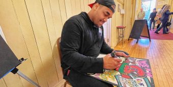 photo of a man seated in a well-lit entry way of a building. he's drawing with markers on a black board on his lap.