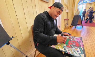 photo of a man seated in a well-lit entry way of a building. he's drawing with markers on a black board on his lap.