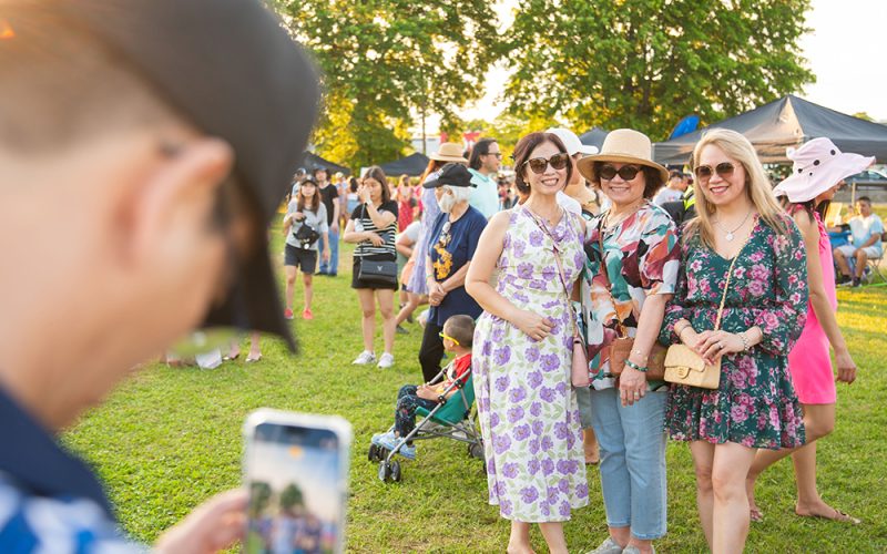 photo of a person holding a phone to take a picture of 3 smiling friends attending an outdoor festival