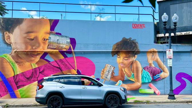 brightly colored mural depicting 2 boys holding tin cans connected by string to their ears. One is also drawing with sidewalk chalk in his free hand.