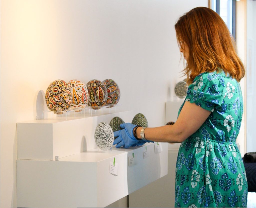 museum worker wearing blue gloves is adjusting a row of intricately painted eggs or pysanka.