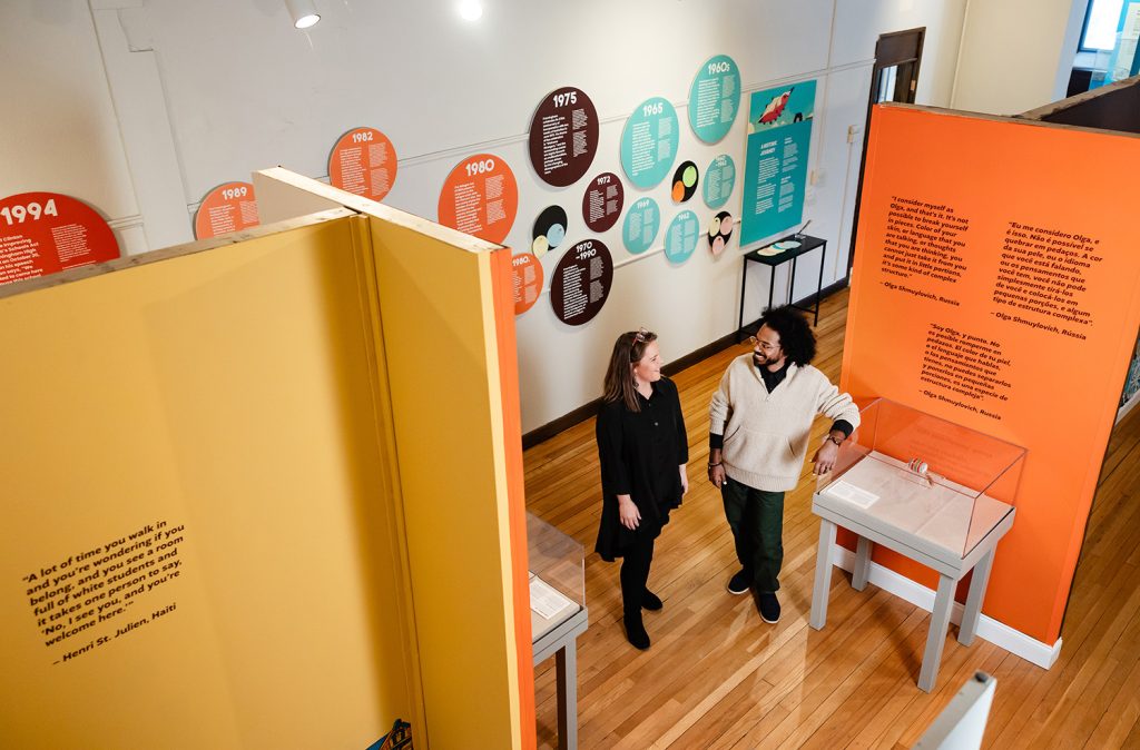 photo of the inside of a gallery. along the back wall is a timeline made of brightly colored circles. in the foreground two people are standing and talking.