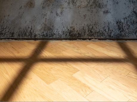 detail of an art installation featuring a metal cube partially suspended over a wooden gallery floor and topped with a crosshatch of wood.