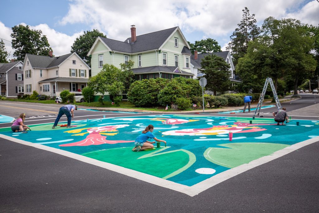photo of people painting a brightly-colored mural covering a large intersection between 2 main roads.