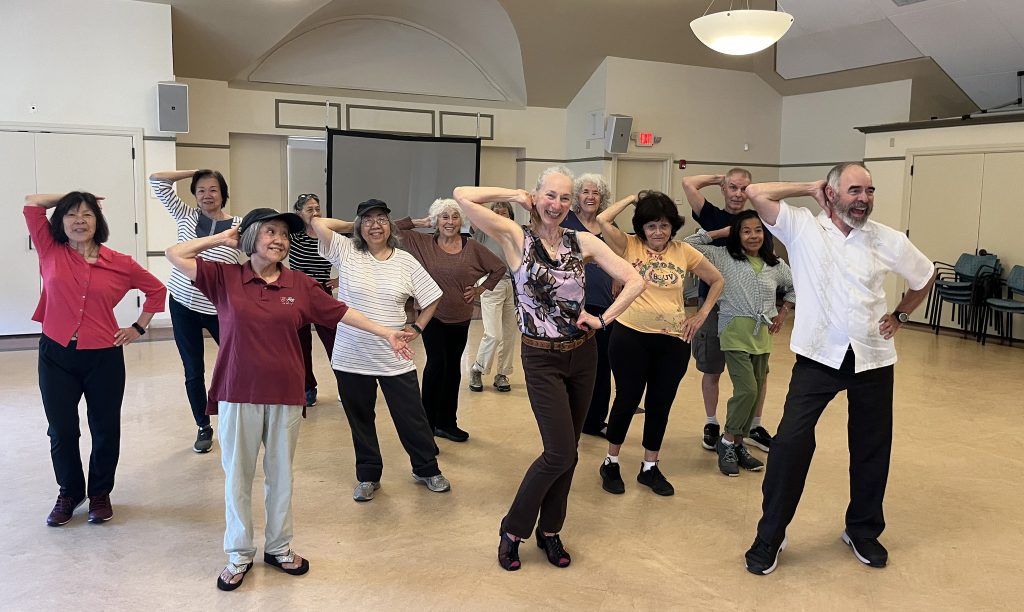 photo of about a dozen older people smiling and dancing in a studio.