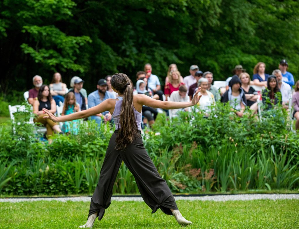 photo of a dancer not facing the camera, their arms extended as they move on the grass outside. in the background the audience looks on.