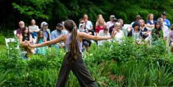 photo of a dancer not facing the camera, their arms extended as they move on the grass outside. in the background the audience looks on.
