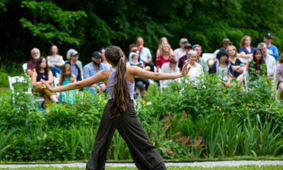 photo of a dancer not facing the camera, their arms extended as they move on the grass outside. in the background the audience looks on.