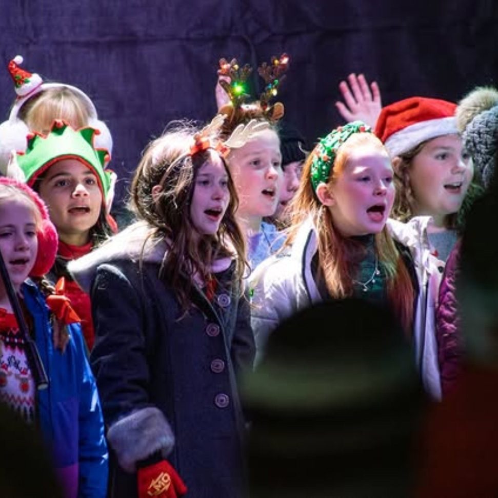 photo of a chorus of children singing, many are wearing festive holiday hats.