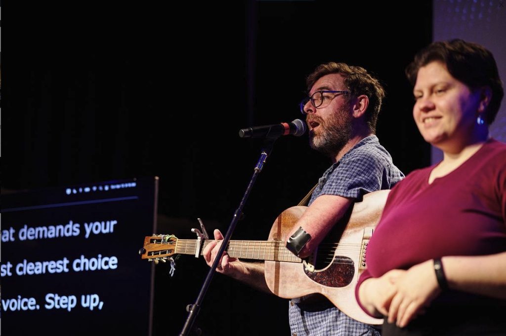 photo of a man playing guitar on a stage. on one side of him stands an ASL interpreter. on the other side of him captions are showing on a large screen for the audience.