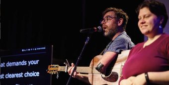 photo of a man playing guitar on a stage. on one side of him stands an ASL interpreter. on the other side of him captions are showing on a large screen for the audience.