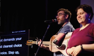 photo of a man playing guitar on a stage. on one side of him stands an ASL interpreter. on the other side of him captions are showing on a large screen for the audience.