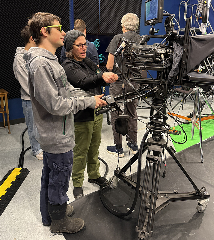 photo of a young person being instructed on how to operate a camera in a television studio by an adult standing next to them.