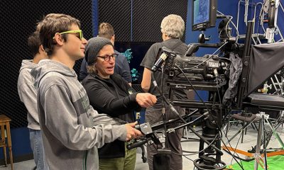 photo of a young person being instructed on how to operate a camera in a television studio by an adult standing next to them.