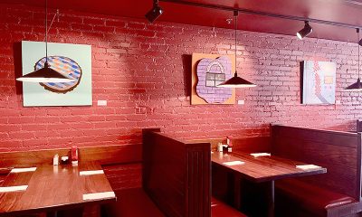 image of the interior of a restaurant with brick walls and wooden booths. Colorful paintings hang on the wall.