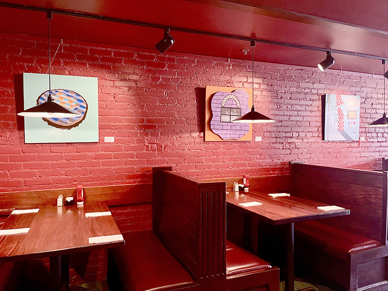 image of the interior of a restaurant with brick walls and wooden booths. Colorful paintings hang on the wall.