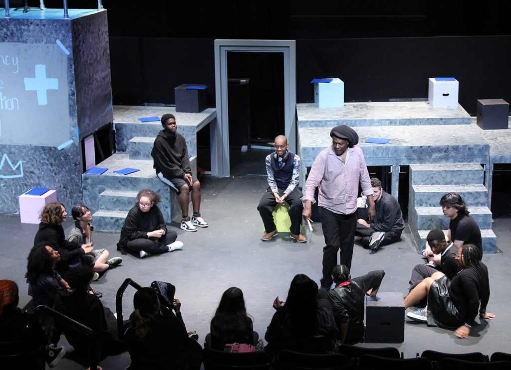 photo of youth theater rehearsal. A man is stands giving direction to a semi circle of actors seated on the floor of a black box theater.