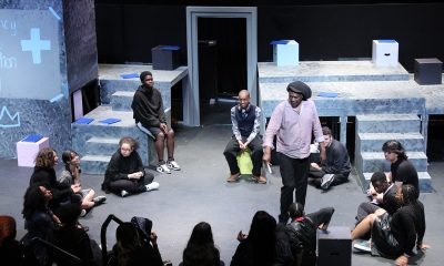 photo of youth theater rehearsal. A man is stands giving direction to a semi circle of actors seated on the floor of a black box theater.
