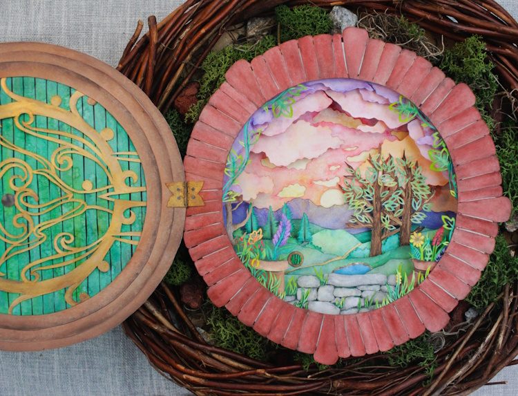 Photo of a vibrantly colored hand-cut paper assemblage that resembles a round door opening onto a peacefully bucolic landscape, sitting within a bird's nest.