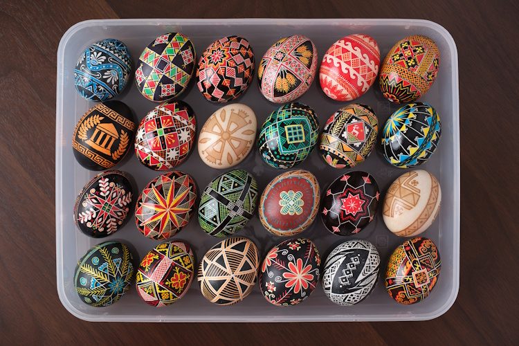 Photograph of eggs with intricate, varied, and colorful designs, neatly arranged in four rows of six.