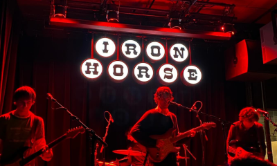 photo of a dark stage only lit by red lights, three musicians playing. Behind them a dramatic black and white sign spells out Iron Horse.
