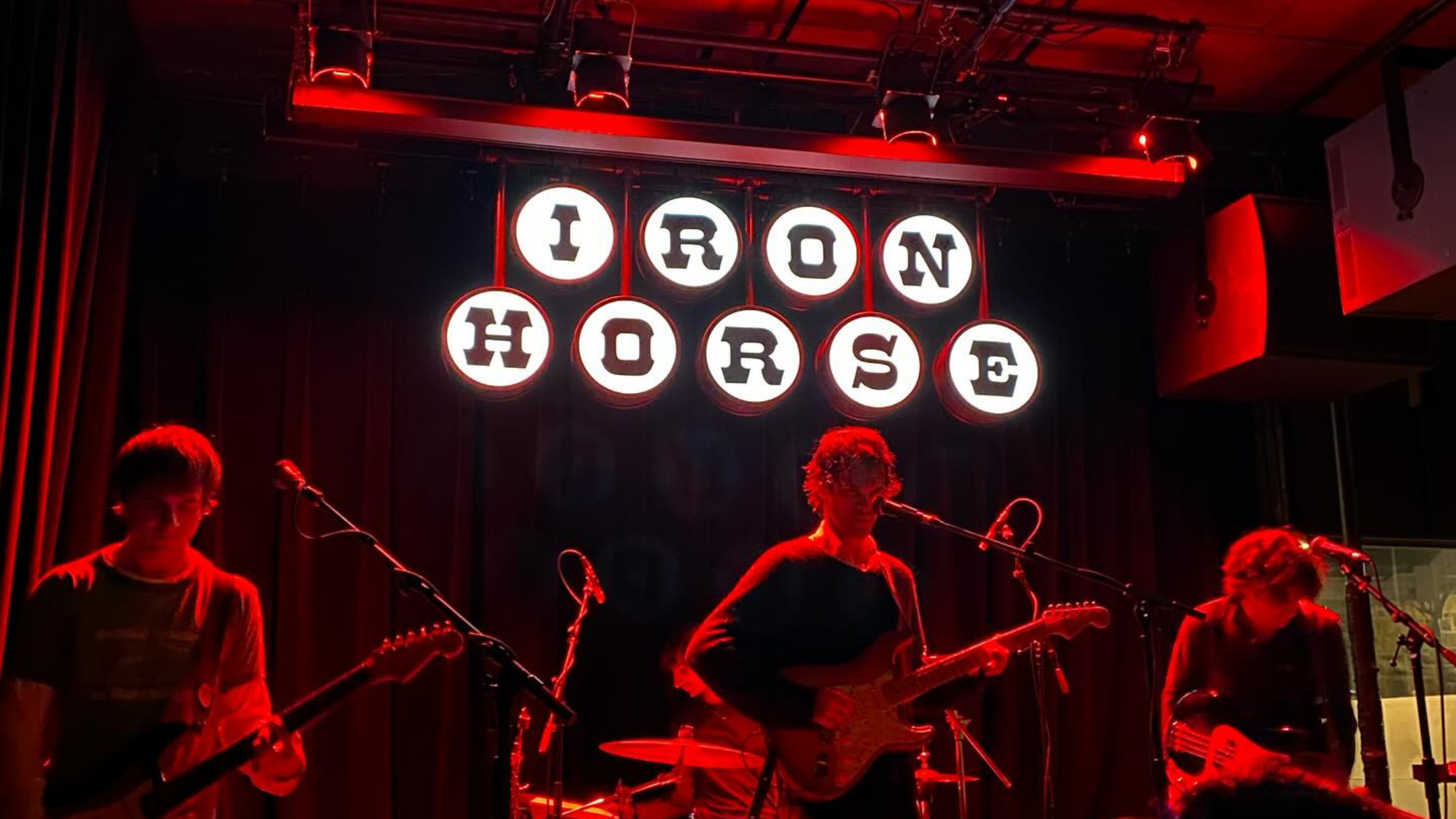 photo of a dark stage only lit by red lights, three musicians playing. Behind them a dramatic black and white sign spells out Iron Horse.