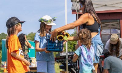 A woman with a streak of blue face paint hands a basket of sunflowers to a young girl wearing a hat adorned with small flowers. It's a sunny day, and the woman and the three children around her are dressed in breezy summer clothing.