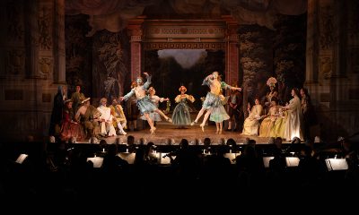 photo of a dark theater with performers dressed in Baroque attire dancing. The musicians playing in front of the stage can be seen in silhouette.