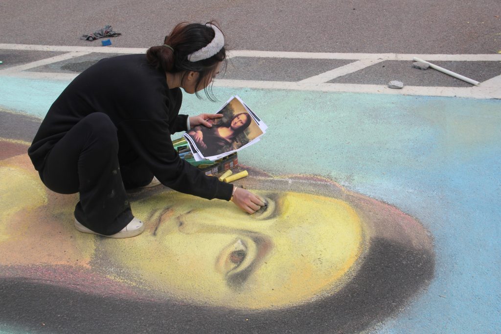 photo of an artist holding a color printout of the painting of the Mona Lisa while crouched down using chalk to draw a rendition of the Mona Lisa in an outdoor parking lot.
