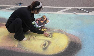 photo of an artist holding a color printout of the painting of the Mona Lisa while crouched down using chalk to draw a rendition of the Mona Lisa in an outdoor parking lot.