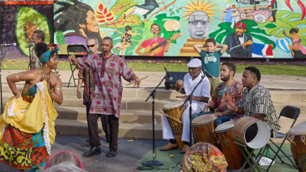 photo of a Latin band performing in front of a brightly painted mural. A dancer wearing a full, yellow skirt performs in front of them.