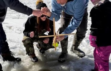detail of a photo of 4 people standing on a frozen lake or pond, looking down at a round hole cut out of it for ice fishing. 2 adults are showing to young people how to use a traditional Nipmuc fishing line.