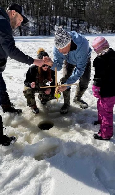 photo of 4 people standing on a frozen lake or pond, looking down at a round hole cut out of it for ice fishing. 2 adults are showing to young people how to use a traditional Nipmuc fishing line.