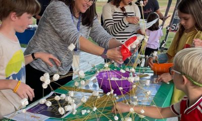 photo of a long table with young people on both sides creating geometric shapes out of marshmallows and uncooked spaghetti noodles.