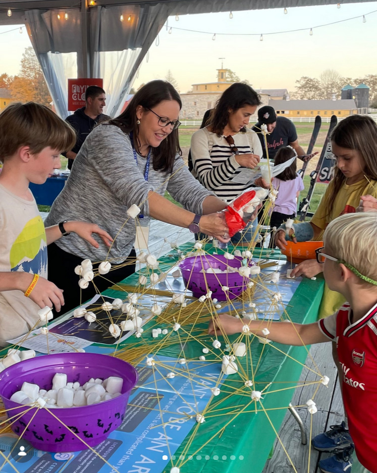 photo of a long table with young people on both sides creating geometric shapes out of marshmallows and uncooked spaghetti noodles.