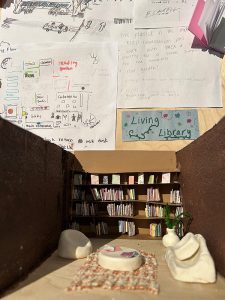 photo of an art piece, a 3-D diorama rendering of a library, including a book shelf, a cozy rug and seating area and plant.