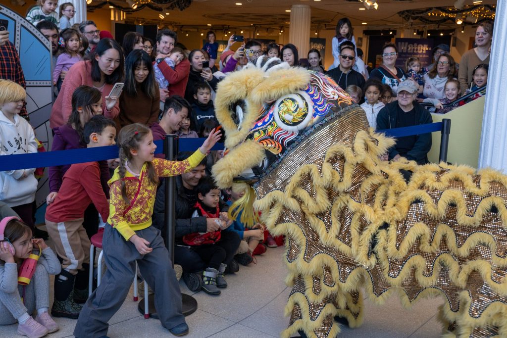 photo of a delighted child reaching out to touch a dragon during a Lunar New Year Celebration. Behind her, a crowd of attendees are smiling and taking photos with their phones.
