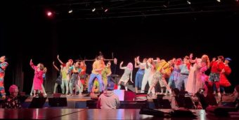 photo from the back of a theater. dozens of people are on stage all facing diagonally in the same direction lurching back in surprise. A handful of musicians are in front of the stage playing in a pit.