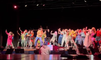 photo from the back of a theater. dozens of people are on stage all facing diagonally in the same direction lurching back in surprise. A handful of musicians are in front of the stage playing in a pit.