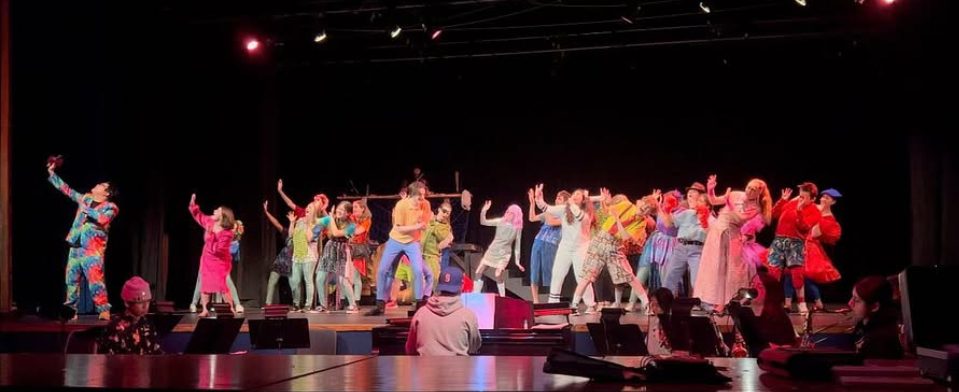 photo from the back of a theater. dozens of people are on stage all facing diagonally in the same direction lurching back in surprise. A handful of musicians are in front of the stage playing in a pit.