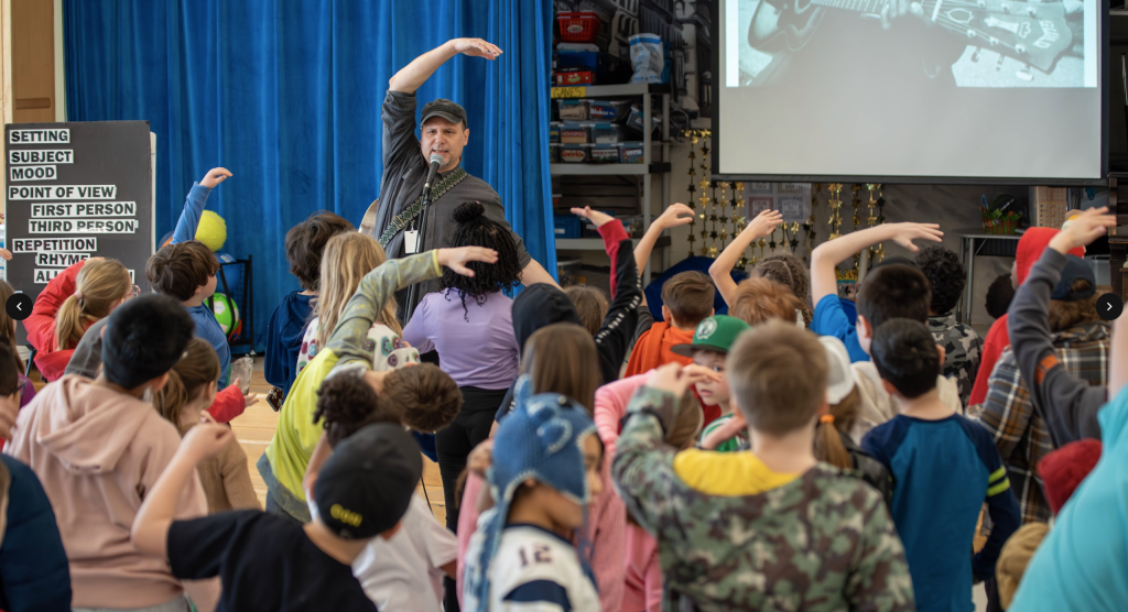 photo of a musician performing in front of a group of school children who are dancing along to his song.