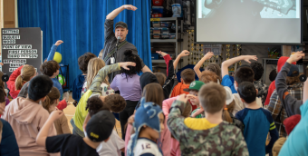 photo of a musician performing in front of a group of school children who are mirroring the way he's gesturing along with the song he's singing.
