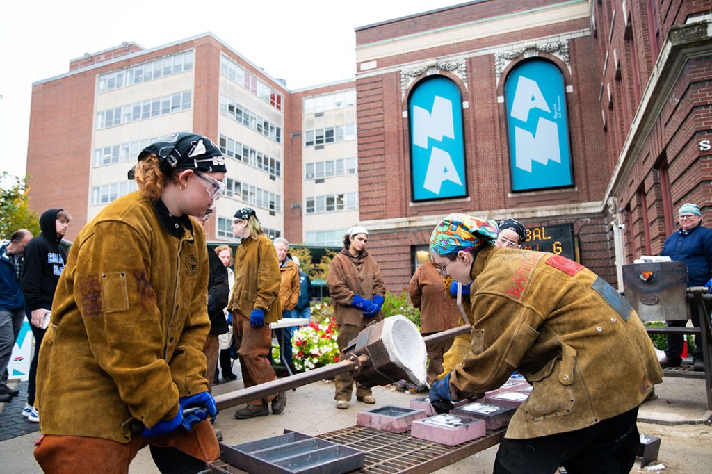 photo of 2 artists pouring liquid metal into molds outside at a public art making event.