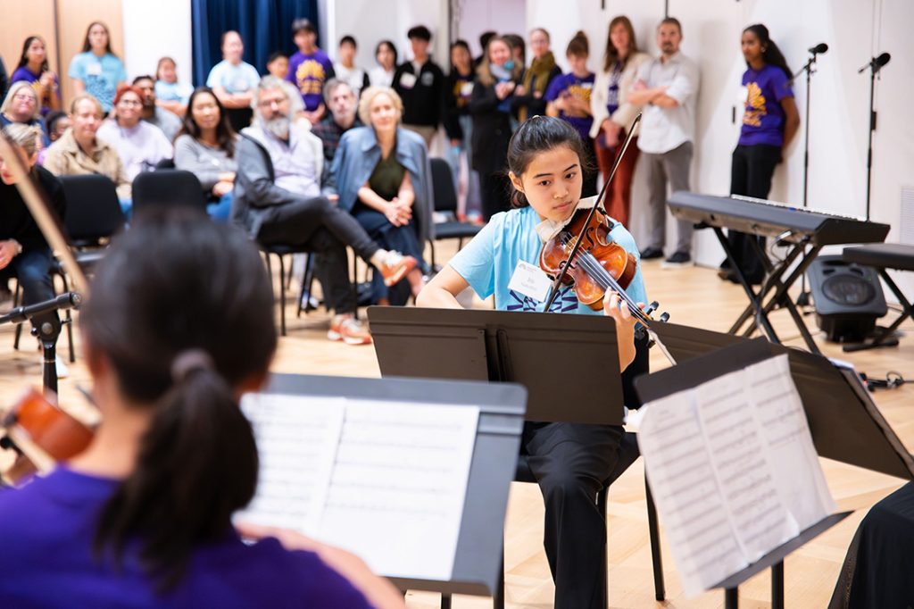 photo of a youth orchestra performing, in the back ground a crowd listens.