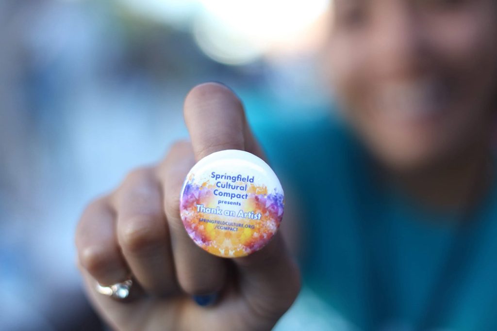 photo of a hand in the background holding a small button with text Thank an Artist in the foreground.