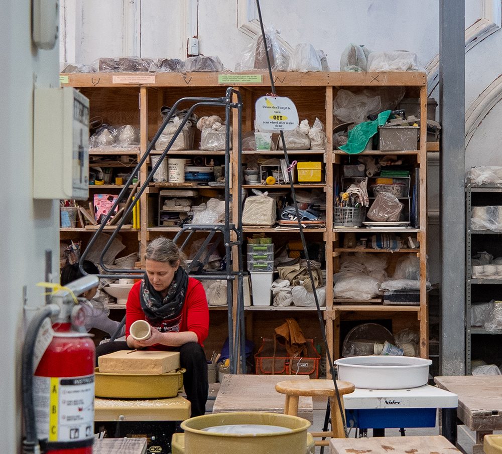 photo of a ceramics studio, a wall of supplies behind an artist working at the wheel.