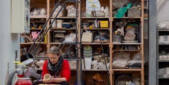 photo of a ceramics studio, a wall of supplies behind an artist working at the wheel.