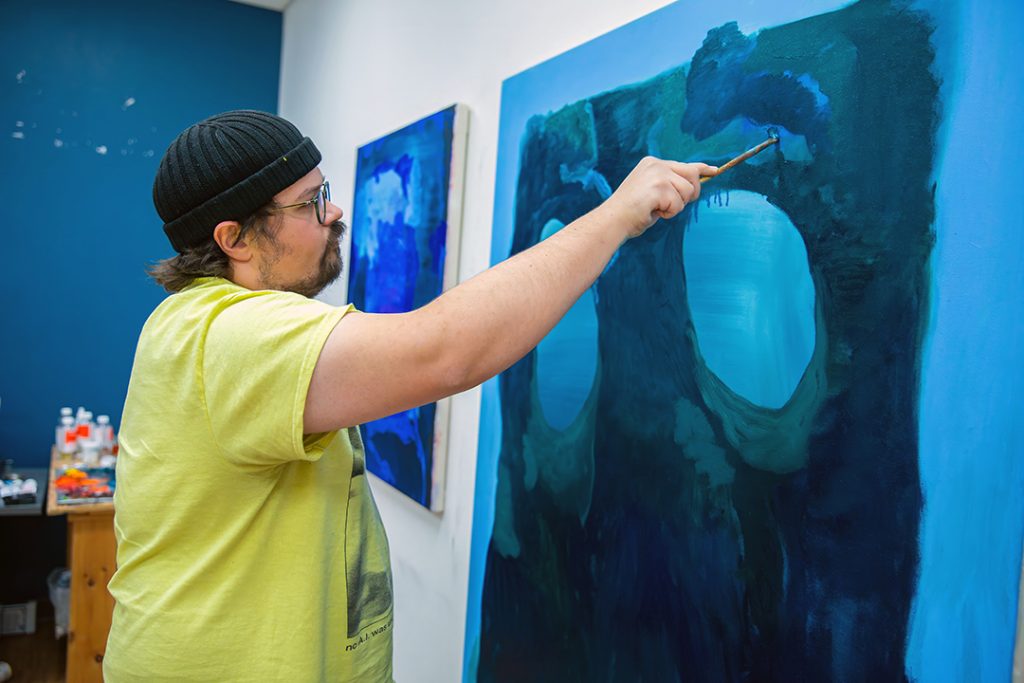 photo of an artist standing, arm extending brushing blue paint on a canvas with many abstract shapes in shades of blue.