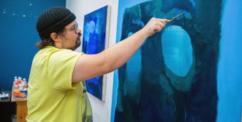 photo of an artist standing, arm extending brushing blue paint on a canvas with many abstract shapes in shades of blue.